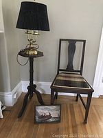 Photo of brass lamp with black pleated shade on octagonal wooden table beside Queen Ann style wooden chair with plaid upholstered seat