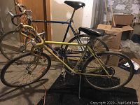 Front side view of two Schwinn racing bicycles together showing yellow Varsity and blue Continental II, placed indoors on concrete floor with boxes in the background.