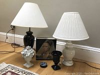 Two ceramic lamps, one black on decorative brass with white pique shade, one white ceramic ginger jar style with pleated shade. Black ceramic vase beside a white lidded Capodimonte-style vase with floral topper, four black Inuit-themed coasters, and National Gallery of Art book.