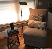 Picture of light beige chenille slipper chair with two patterned pillows and rust red wooden side table holding stacked books and a pottery vase in front of window with blinds.