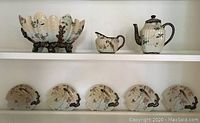 Full view of tea set pieces displayed on shelf including teapot, pitcher, bowl, and five decorative plates with scalloped edges and gilded handles.