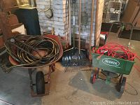 Photo showing red True Temper USA wheelbarrow loaded with coiled garden hoses, Scotts seed spreader with extension cord, and two garden rakes leaning behind.