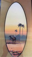 Oval-shaped watercolor painting of an Egyptian desert landscape featuring a camel with a rider, palm trees, and pyramids under a soft sunset sky. Framed under glass with a tan mat in a black rectangular frame.