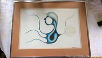 Framed Indigenous art print featuring abstract profile figure with flowing lines in blue, black, and gray tones on white background, matted and under glass on light brown mat board.