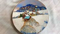 Full view of round hand painted enamel plate showing winter village scene with snow and houses.