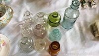 Nine antique glass ink wells in clear, green, blue glass and one taller, amber embossed glass bottle on a white tablecloth.