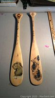 Two wooden paddles side by side showing painted animal designs and paw prints with ruler for scale