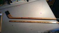Full view of wood hockey stick alongside a ruler for scale, showing natural wood, black tape on blade and branding on shaft.