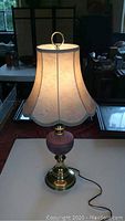 Full view of the table lamp showing brass base, pink art glass section, and cream floral fabric shade with scalloped edges and braided trim.