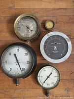 Five vintage gauges laid out on a wooden surface revealing various sizes and styles including large black and white pressure gauge, wind speed gauge, and smaller dials.