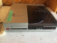 Front view of Aiwa LX-70 turntable showing power button, linear tracking program buttons, and dust cover. Turntable placed on a white wooden surface with some wear marks.