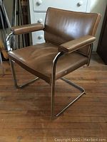 Front angle showing chrome frame, caramel brown leather upholstery, padded armrests, and seat cushion with normal wear.