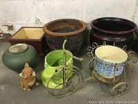 Photo showing all garden pots and decorative planters grouped on floor: large clay pot, square pot, green pot, ceramic boot, fox figurine, and two metal planter carts.