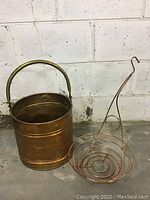 Copper pail and copper wire plant hanger shown together on concrete floor against a white wall.