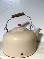 Front angled view of the antique cream-colored enamel kettle showing wooden handle and red lid knob with base wear.