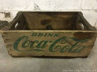 Front-facing photo of the rectangular worn wooden crate showing green 'Drink Coca-Cola' branding on the side.