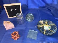 Full lot display of four glass decorative plates, rose quartz crystal, glass jar with lid, glass paperweight, four textured glass coasters, and orange glass sculpture arranged on blue fabric.