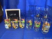Photo showing six Muppets cocktail glasses and two Hard Rock Orlando hurricane glasses arranged on blue cloth with lot 23 sign