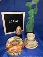 View of all four items: marble style stone bowl with lid, stone ornament bowl, painted stone egg on wooden base, and green plastic woman sculpture holding a fan, against blue background with letterboard showing 'LOT 31'.