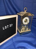 Front angle view of the gold colored quartz carriage clock with clear glass panels showing dial and decorative top ring handle.