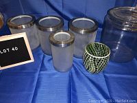 View of four frosted glass containers with silver lids, clear glass jar with lid, and green glass mosaic pot on blue cloth.