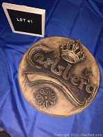 Top angled photo showing the entire round wooden Carlsberg sign with carved logo and crown resting on a blue cloth background next to a small black and white sign labeled 'LOT 41'.