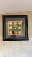 Full view of black framed plaque mounted on wall showing nine gold-colored coins with Coca Cola Olympic logo center