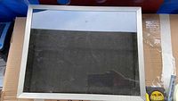 White shadow box frame with glass front, showing surface marks and smudges, placed on cardboard.