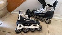 Pair of Bauer Tuik Performance One Up Supreme Ascendant men's inline rollerblades, black and silver with white laces, size 8, placed on the floor near a wall.