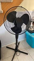 Photo of a freestanding black plastic 16 inch blade pedestal fan, showing the full unit against a wall next to stairs.