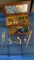 Photo of open wooden jewelry box showing multiple compartments holding silver and gold-tone jewelry including earrings, rings, and bangles, plus watches, and lip gloss tubes