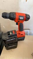 Black & Decker 12V cordless drill with two batteries resting on a light wooden surface, plugged wall socket behind.