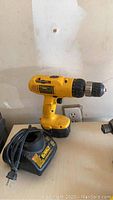 Yellow DeWalt cordless drill and charger placed on a table with a plain wall background, showing the drill standing upright next to its charger with cord coiled.