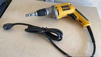 Single photo of a yellow and black DeWalt drywall screwdriver with long power cord placed on a wooden surface.