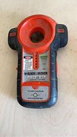 Photo of a Black and Decker Bulls Eye stud finder placed on a wooden surface. The device is black with orange-red detailing and visible labels, showing signs of use.
