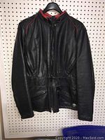 Full front view of black leather woman's motorcycle jacket with red piping, hanging on a hanger against a pegboard.