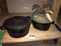 Photo showing both the 10-inch cast iron Dutch oven and the 8-inch cast iron base with the colorful ceramic dragon-shaped lid side by side on a wooden shelf.