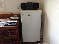 Magic Chef bar fridge closed with RCA stereo on top, viewed from front showing wear and proximity to wall and furniture