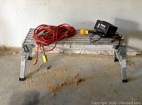 Aluminum scaffold bench supporting battery charger and extension cord, on concrete floor with sawdust.