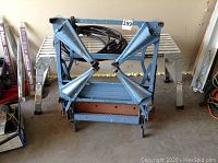 Blue folding scaffold frame in folded position with attached cable coiled inside.