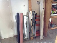 Photo showing 2 metal sawhorses, black and pink vapour barrier rolls, roll of heavy paper, and shelving sections against wall