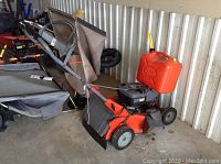 Side view of red steel deck lawnmower with grass bag and gas can alongside