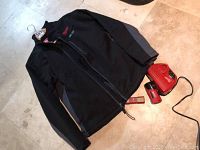 Full view of black Milwaukee heated jacket laid flat with red battery and charger to the side