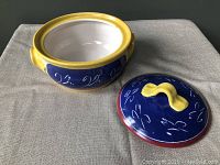 Serving bowl with lid removed shown on fabric surface, blue with white script and yellow trimming.