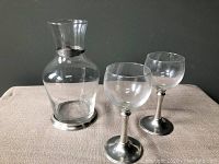 Clear glass decanter and two matching wine glasses with pewter bases and trim set on table against dark background.