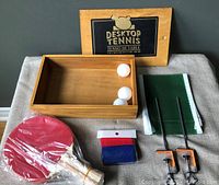 Wooden box, two red paddles, two balls, green net with clamps, and fabric strip on grey surface