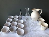 Set showing 12 white ceramic ramekins on left, baster in center, clear glass decanter behind baster, clear sherry glasses in front, and large white ceramic water jug on right