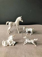 Four white horse figurines displayed on beige surface against dark background, including large horse with repaired leg, smaller horse, reclining unicorn with gold horn, and galloping horse.