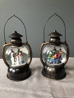 Photo of two snow globes side by side, showing the winter scenes inside and lantern-shaped bases with metal handles.