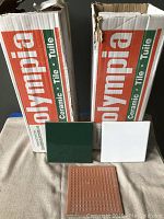 Two Olympia branded boxes containing ceramic tiles with one green tile and one white tile placed upright in front.